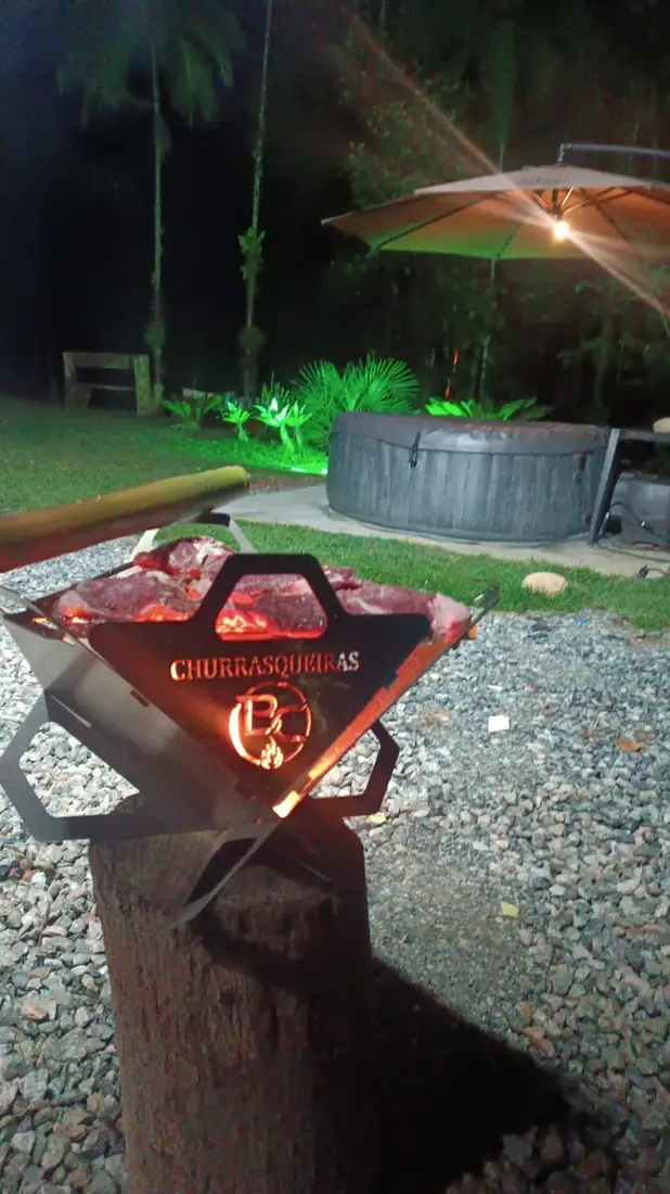 Churrasqueira B.C ONYX acesa à noite, com a brasa iluminando o logo cortado a laser na chapa de inox
